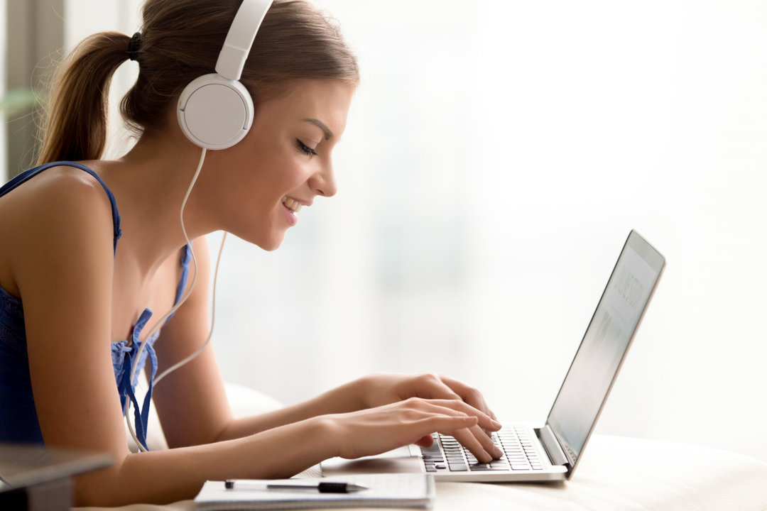 Side view portrait of smiling young woman in headphones typing on laptop while taking online course or personal training for self-education, qualification increase. Happy lady studying in Internet