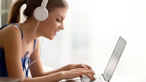 Side view portrait of smiling young woman in headphones typing on laptop while taking online course or personal training for self-education, qualification increase. Happy lady studying in Internet