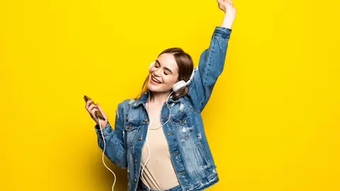 Happy cheerful woman wearing wireless headphones listening to music from smartphone studio shot isolated on yellow background
