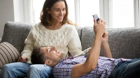 Smiling happy millennial couple using smartphone relaxing on couch, boyfriend and girlfriend checking social network news online on cell together at home, young man showing woman new mobile phone app